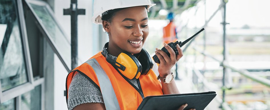 Woman with orange work vest, hard hat and headset looking at screen and holding comms device