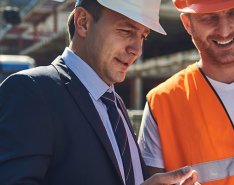 Man in orange vest and hard hat talks to man in suit and hard hat