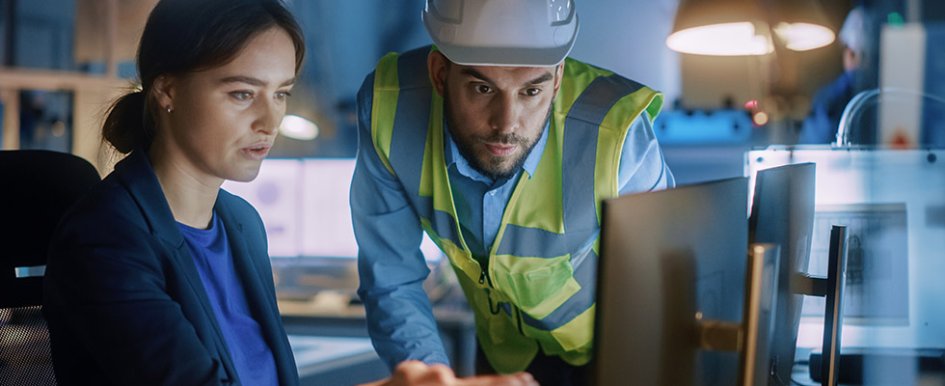 Woman in business attire on computer while man in vest and hard hat looks at screen