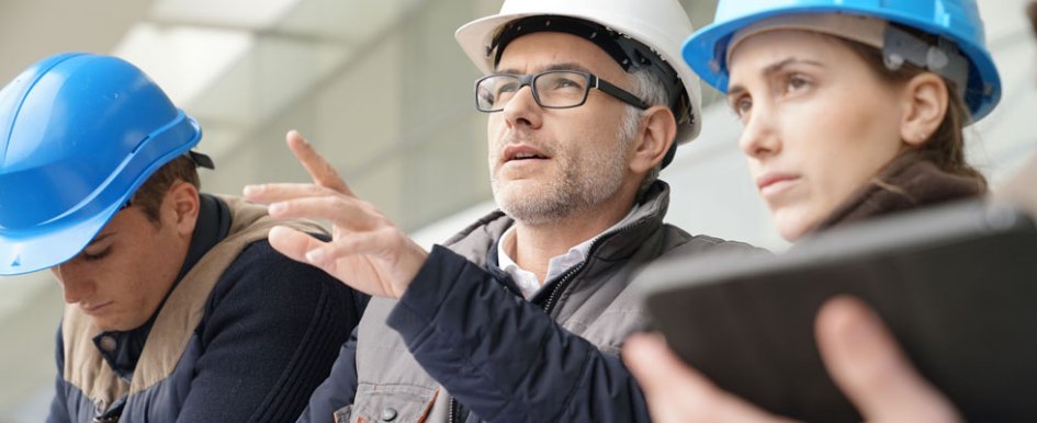 Two people wearing hardhats look forward. One of the people is wearing glasses and pointing at something out of frame. A third person, also wearing a hard hat, looks down.